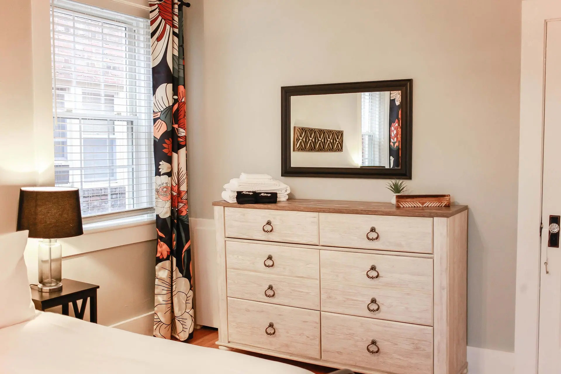 A dresser with a mirror above it, a lamp on a nightstand, and a curtain with a floral pattern.