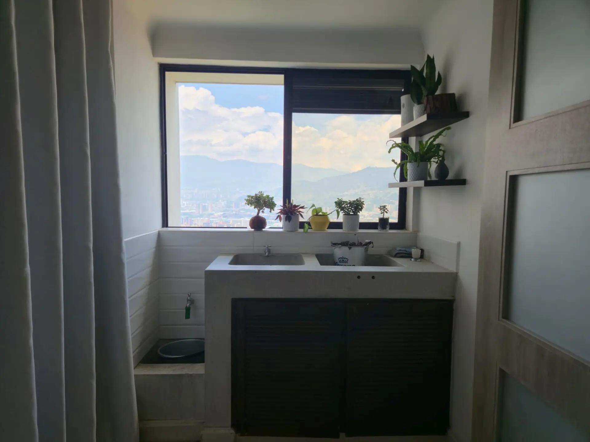 A sink and plants are in a room with a view of mountains and a city.