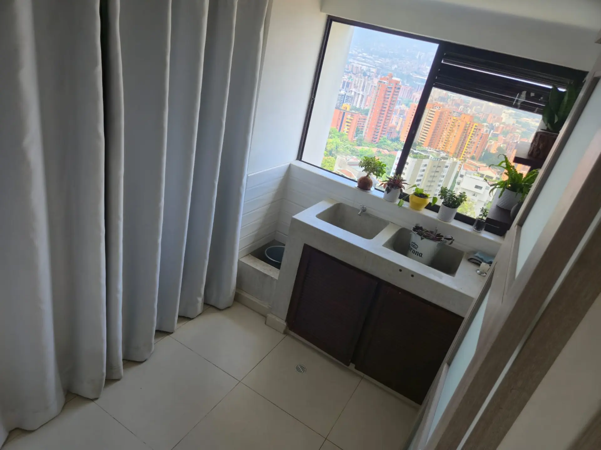 A double sink with cabinets below and plants on the windowsill overlooking a city.