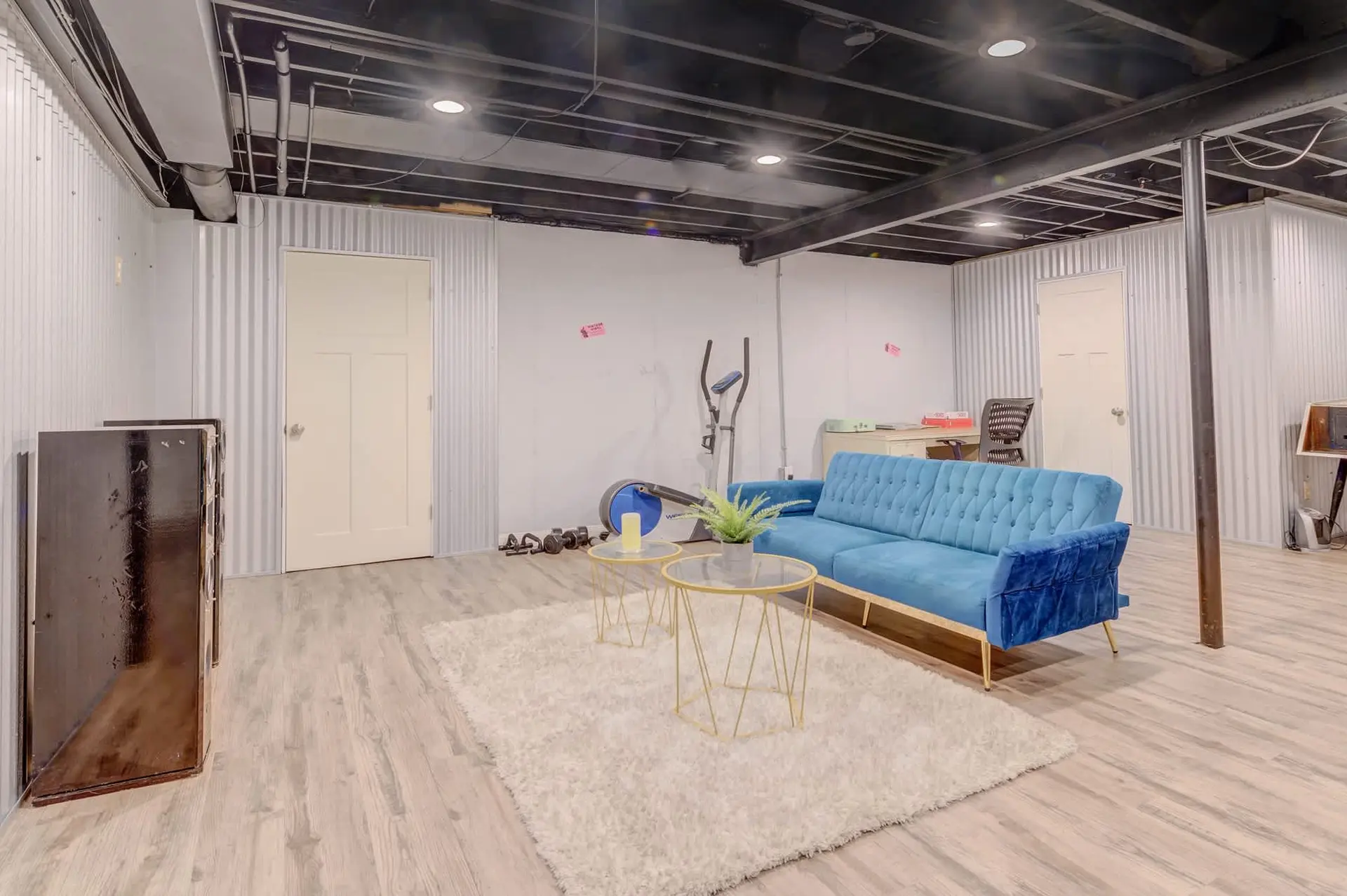 A modern, finished basement with a blue couch, elliptical machine, and corrugated metal walls.