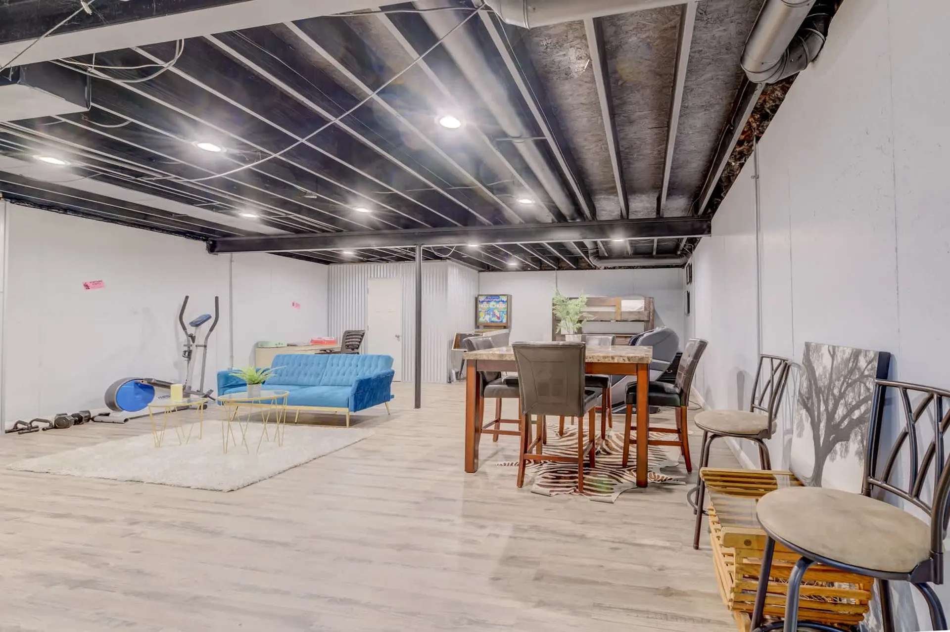 A basement living area with an elliptical, blue couch, dining table, and chairs.
