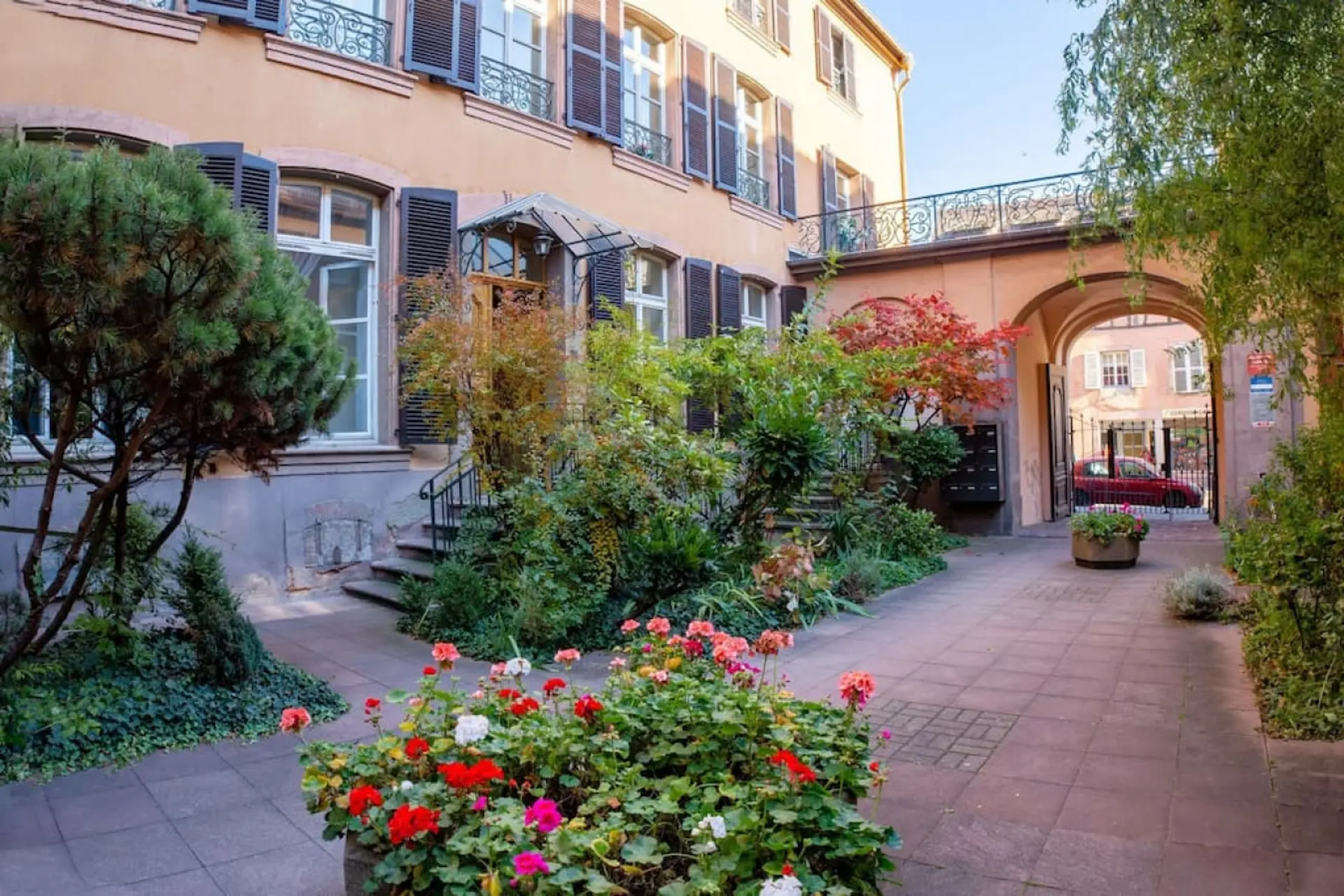 A charming courtyard with colorful flowers, lush greenery, and an arched walkway leading to a street