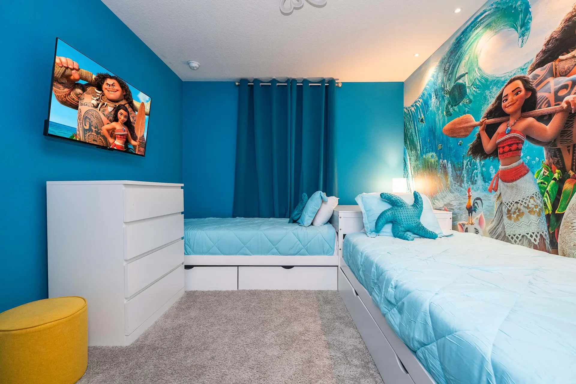A bedroom with blue walls, a Moana mural, two beds with blue bedding, and a dresser.