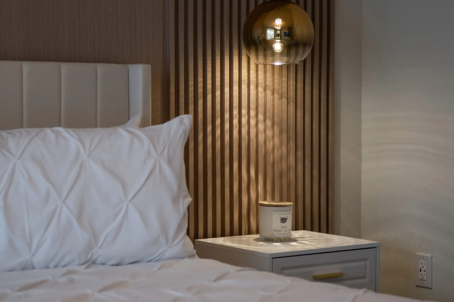 A bedside table with a candle is next to a bed with a white headboard.