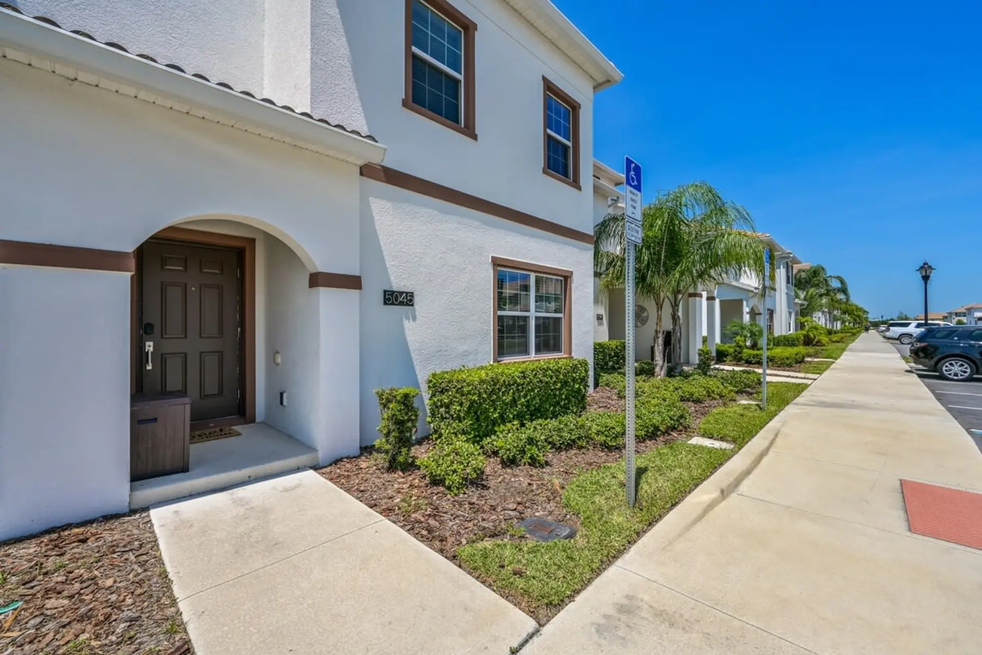 A white townhouse with a brown door and brown trim has a sidewalk leading to it.