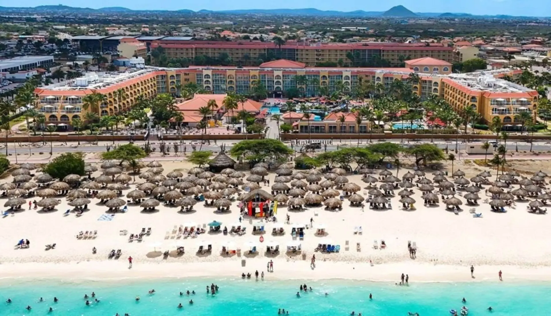 An aerial view of a crowded beach with thatched umbrellas and a resort in the background.