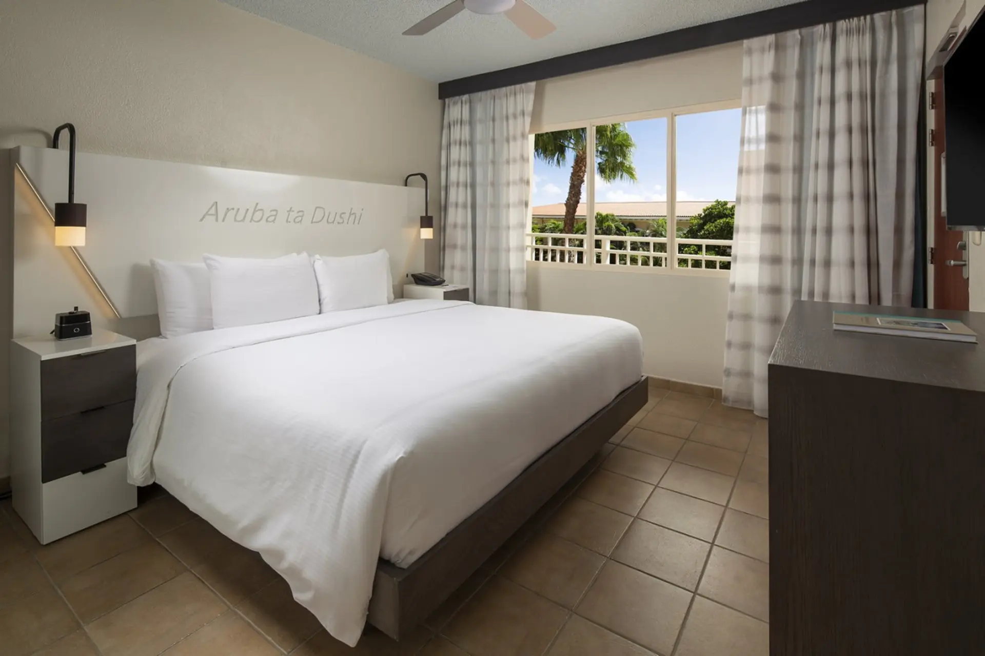A bright, modern hotel room with a king-size bed, white linens, and a view of palm trees.