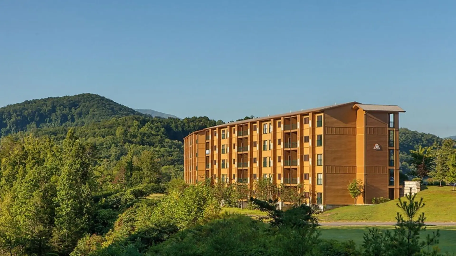 A long, wooden building with balconies stands amidst lush green trees and rolling hills.