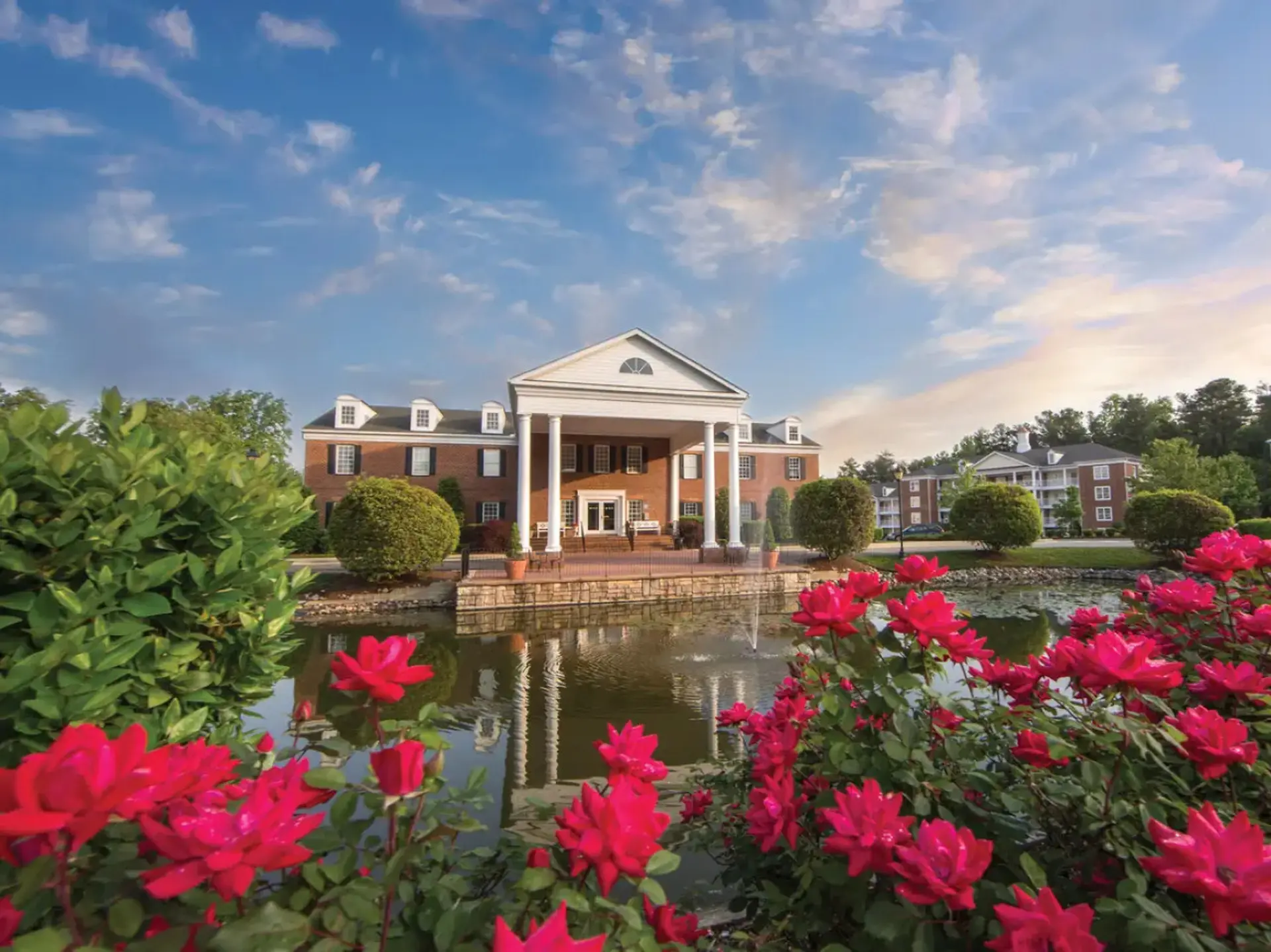 A brick building with a white portico is reflected in a pond, with bright red roses in the foregroun