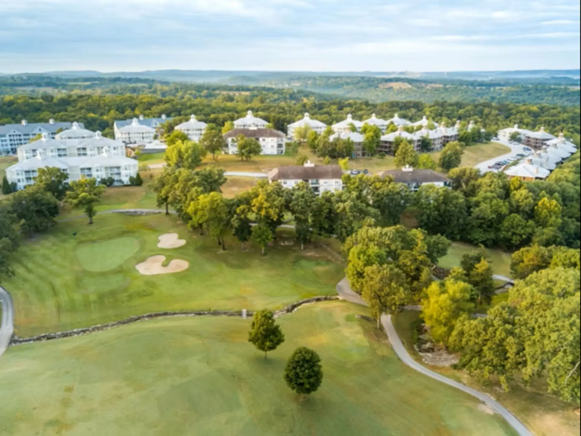 Aerial view of a golf course with resort-style buildings nestled among trees under a cloudy sky.