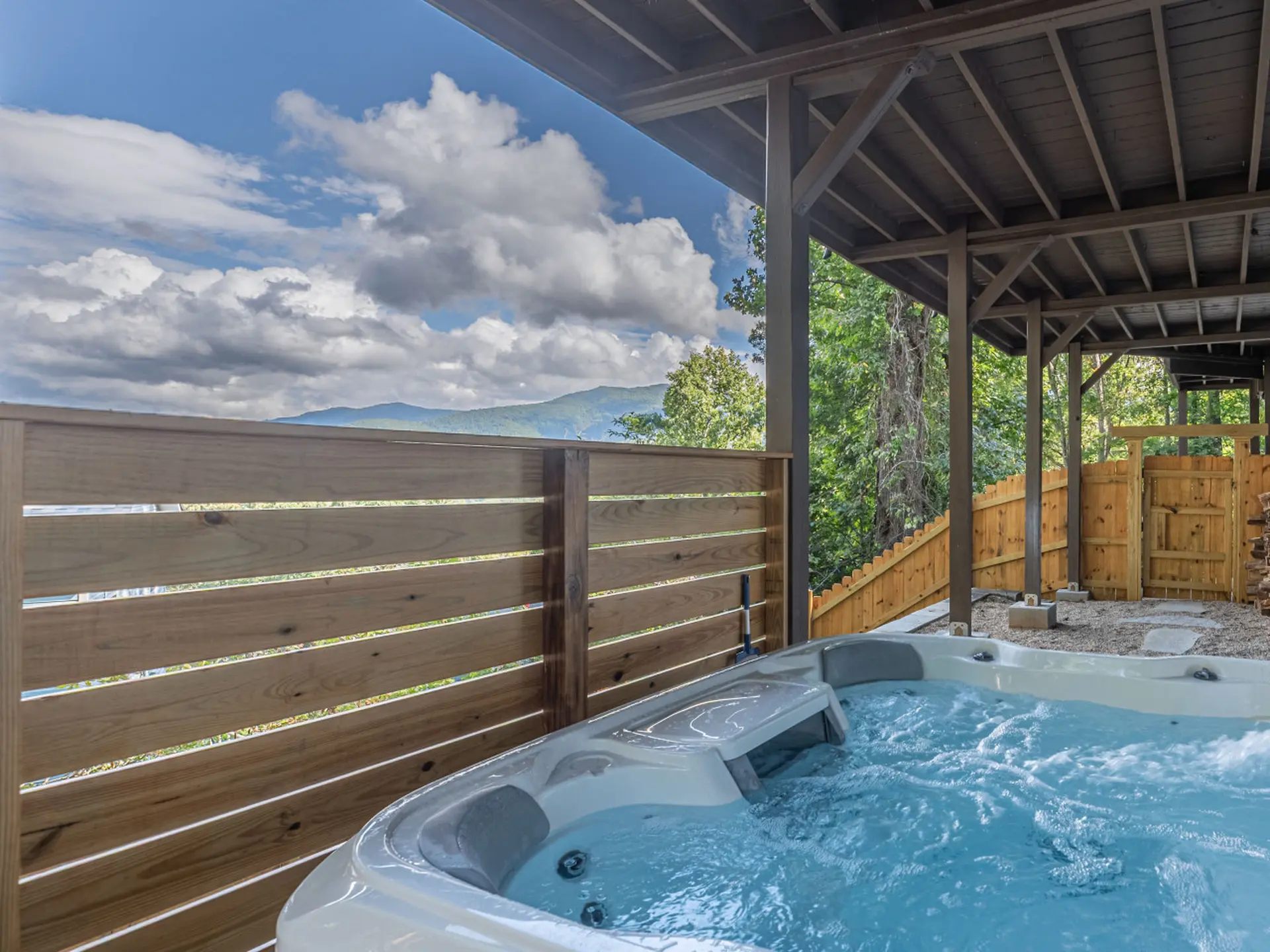 Hot tub on a deck overlooking mountains and trees under a cloudy sky.