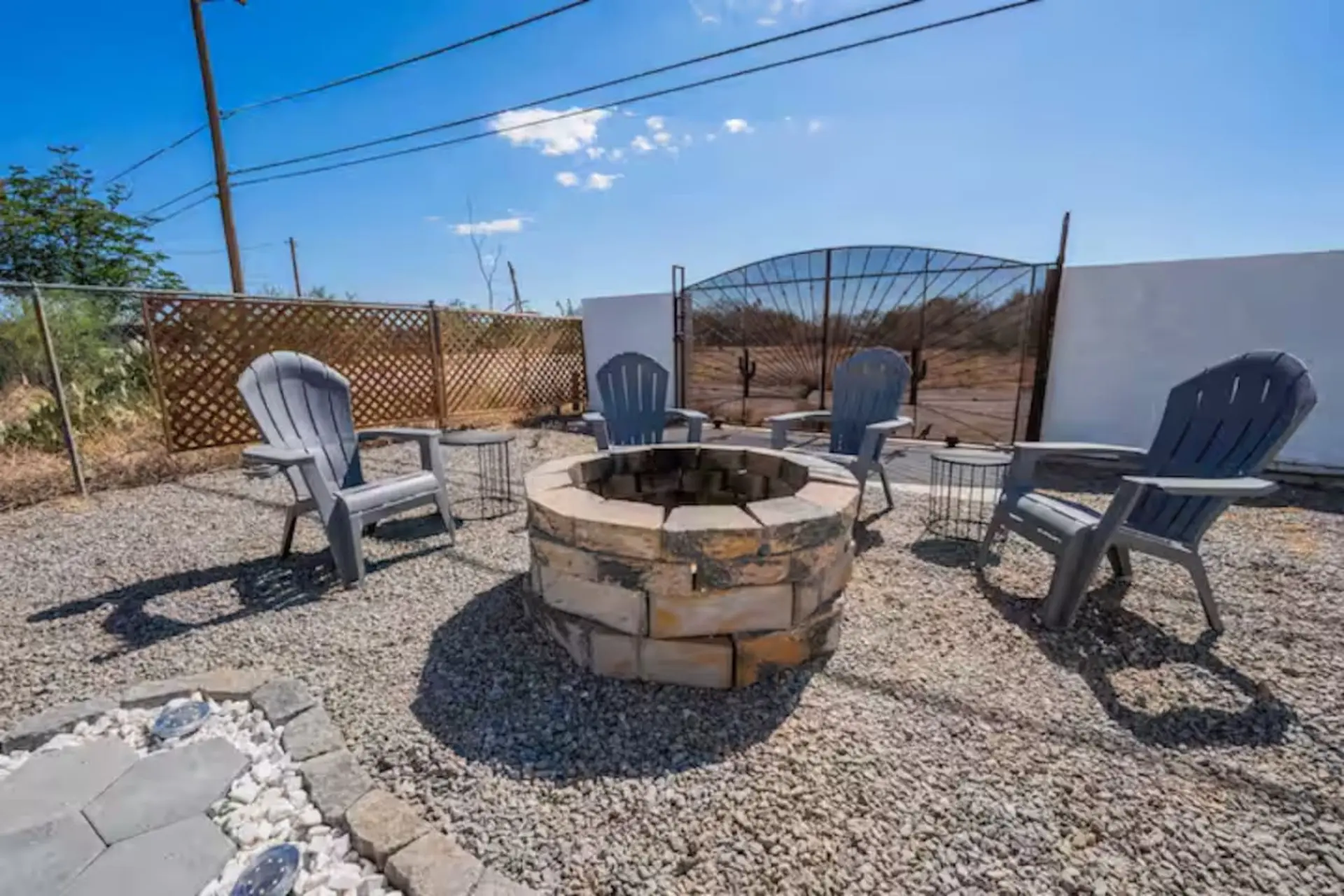 A patio with a stone fire pit, four Adirondack chairs, and a metal gate in the background.