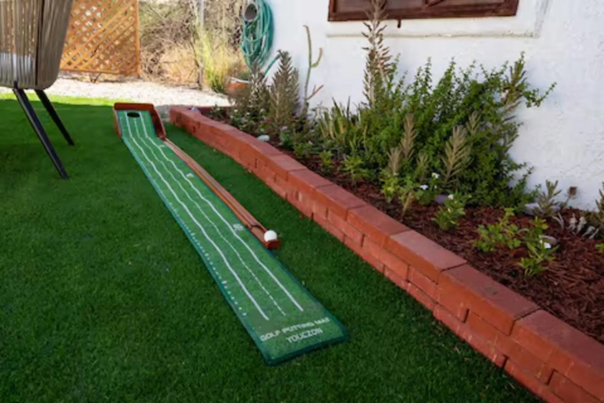 A golf putting mat with a ball sits on artificial grass next to a brick border and garden.