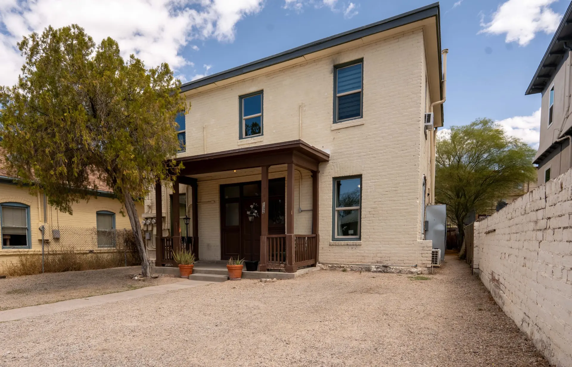 A front view of a light yellow, two-story brick house with a dark brown porch and a tree in front.