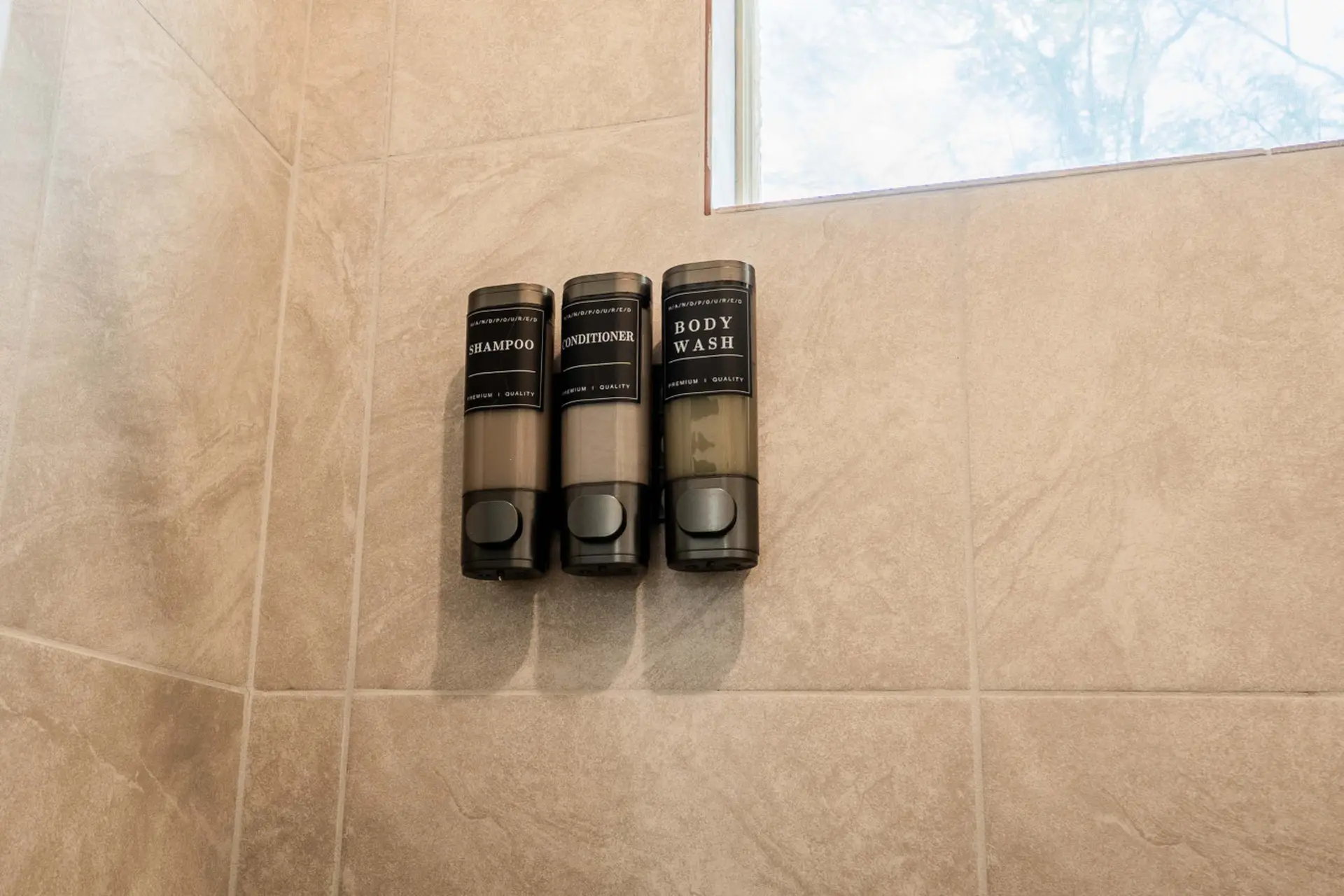 Three clear plastic dispensers with black tops are mounted on a tiled wall in a shower.
