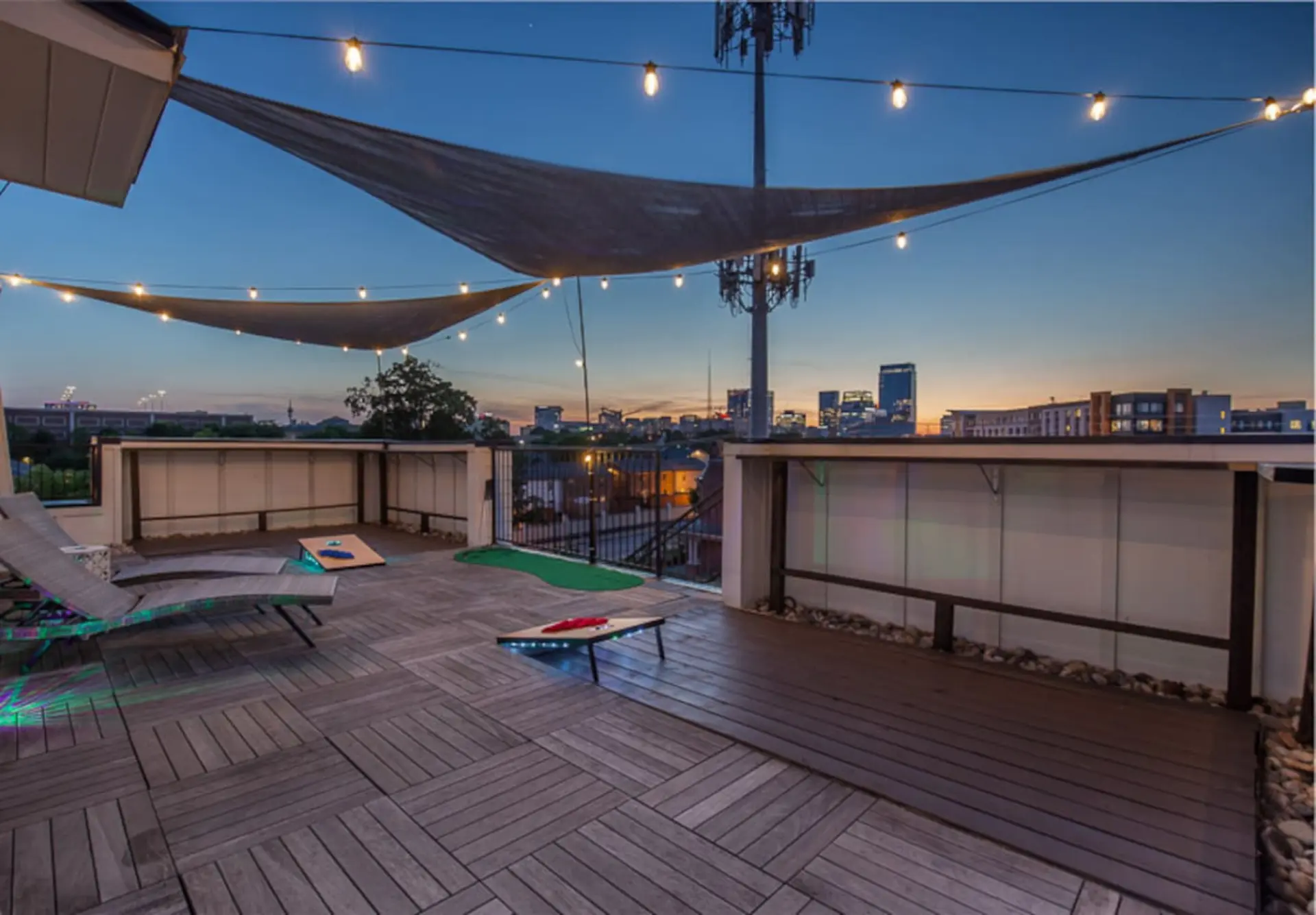 Rooftop deck at dusk with city skyline, string lights, cornhole boards, and lounge chairs.