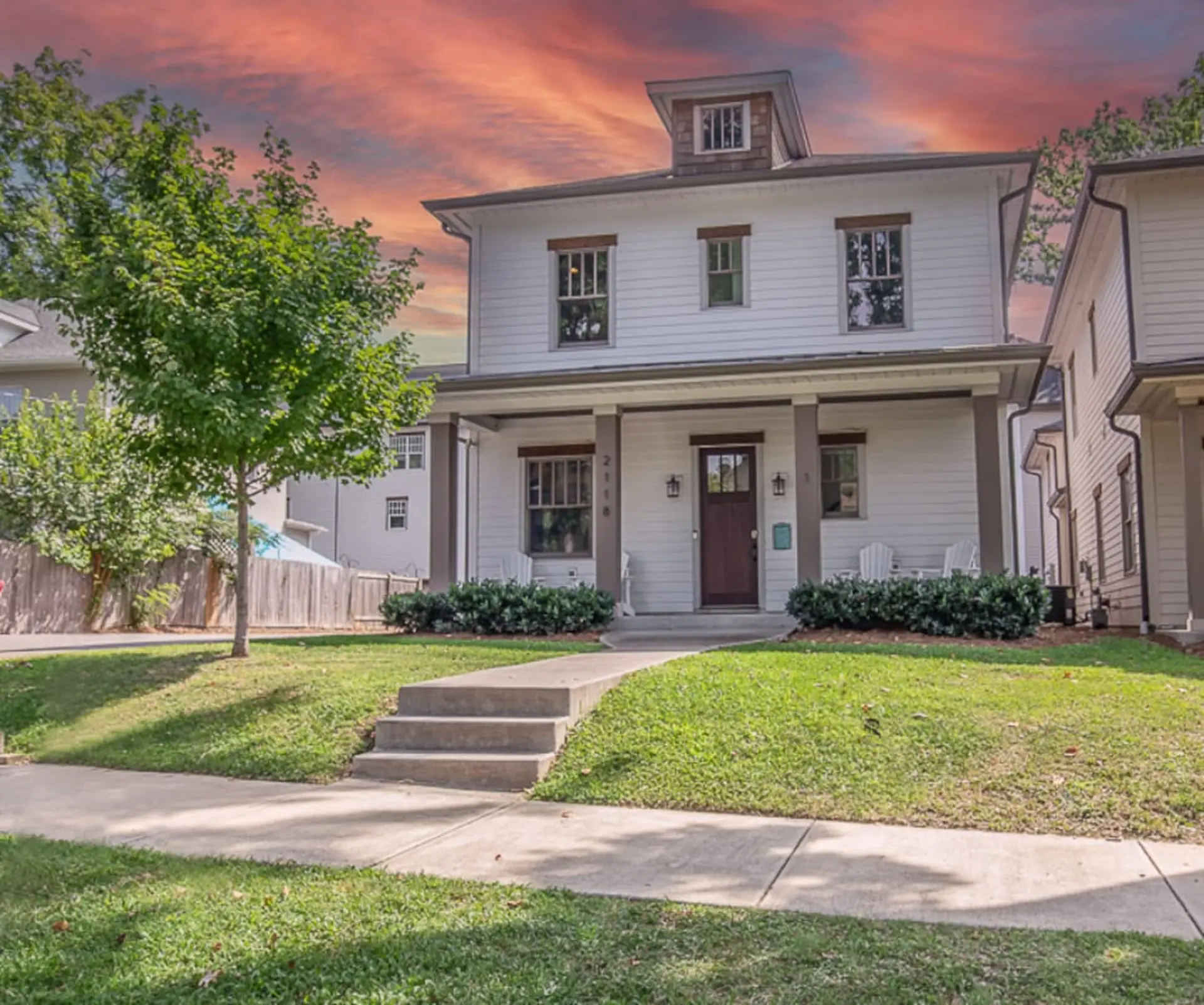 A white, two-story house with a front porch and manicured lawn under a dramatic sunset sky.