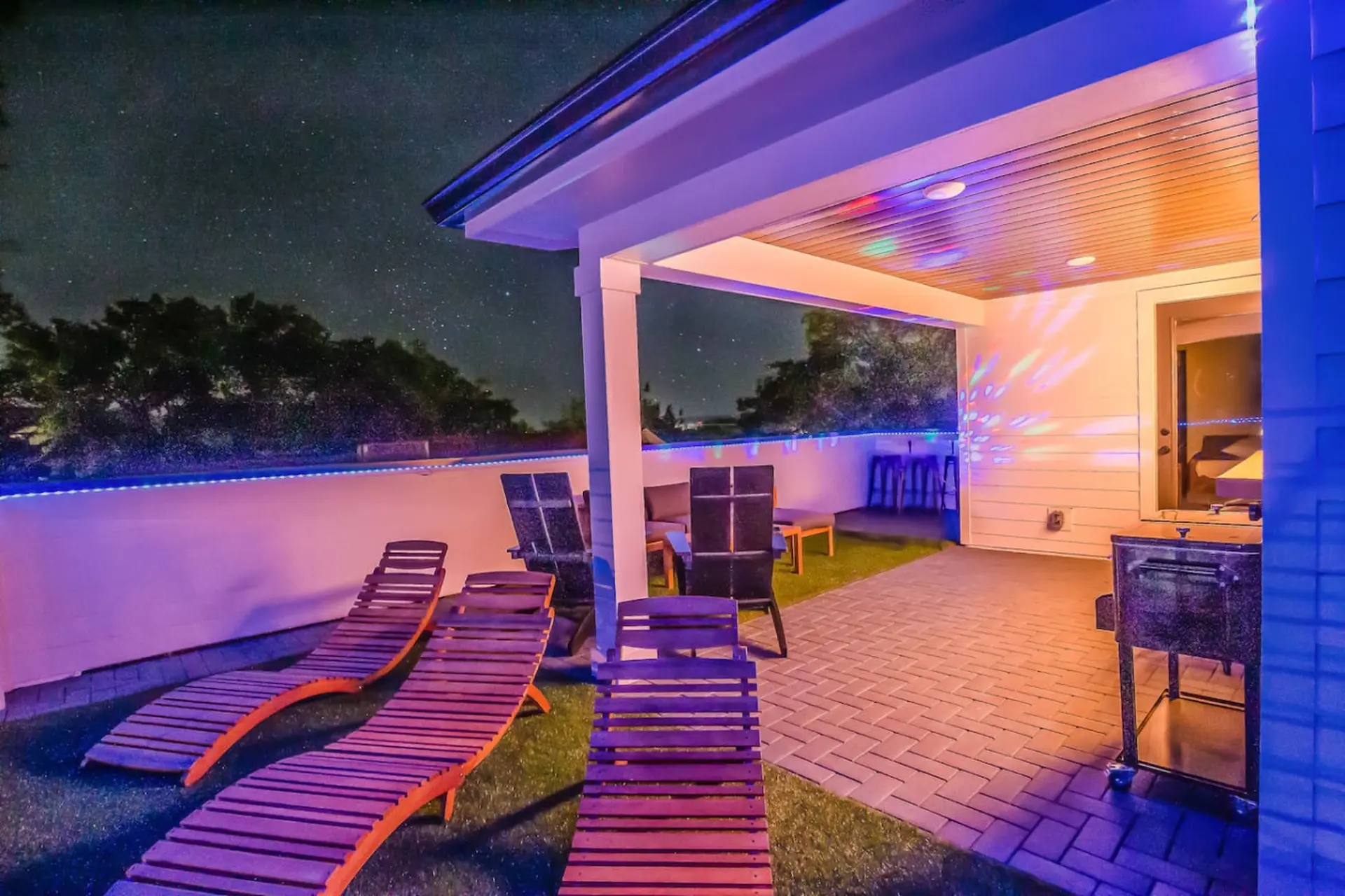 Night view of a patio with lounge chairs, a grill, and colorful lights reflecting on the ceiling.