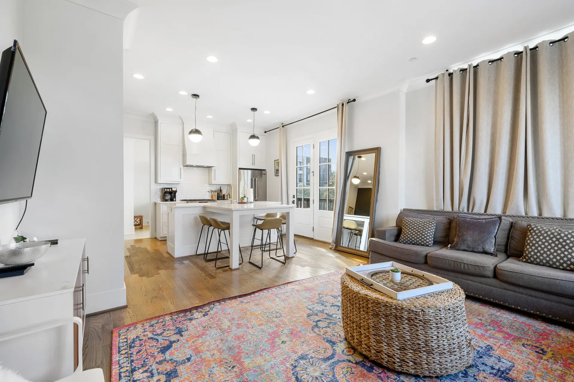 A gray sofa sits on a colorful rug in a bright living room with a kitchen.