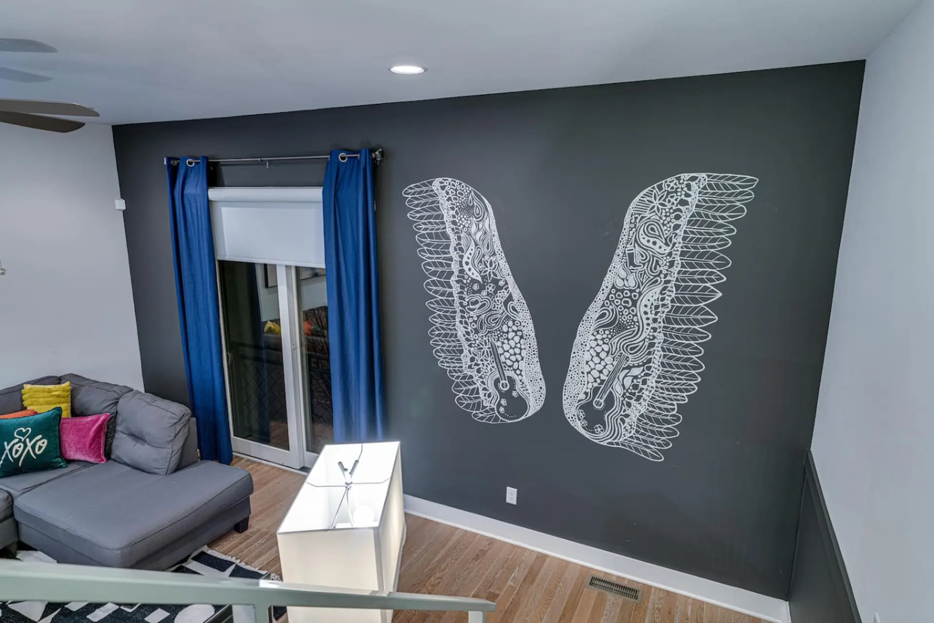 A living room with a gray sectional sofa, bright blue curtains, and a wall mural of white wings.