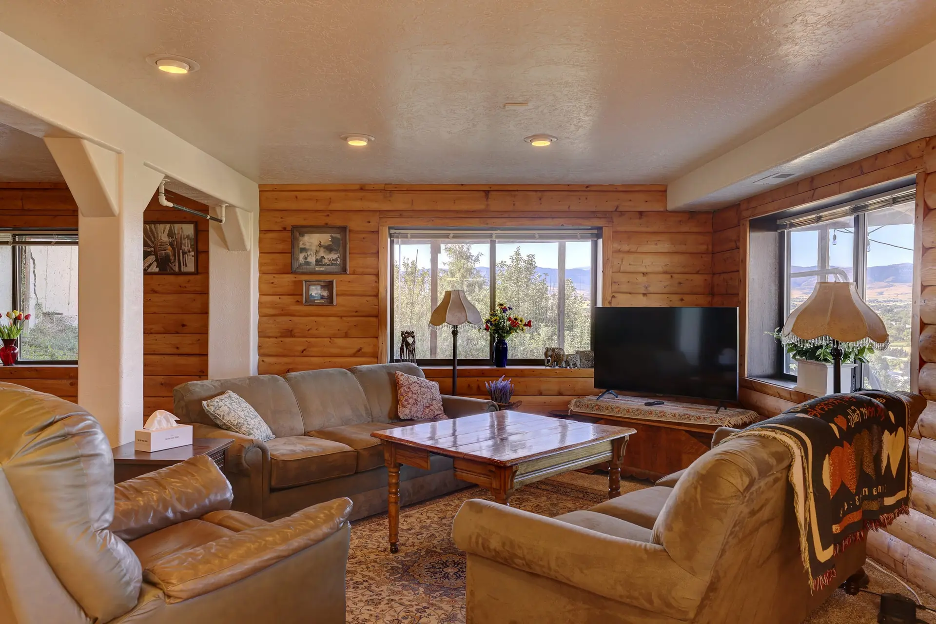 A rustic living room with log walls, plush furniture, and a large window overlooking a scenic landsc