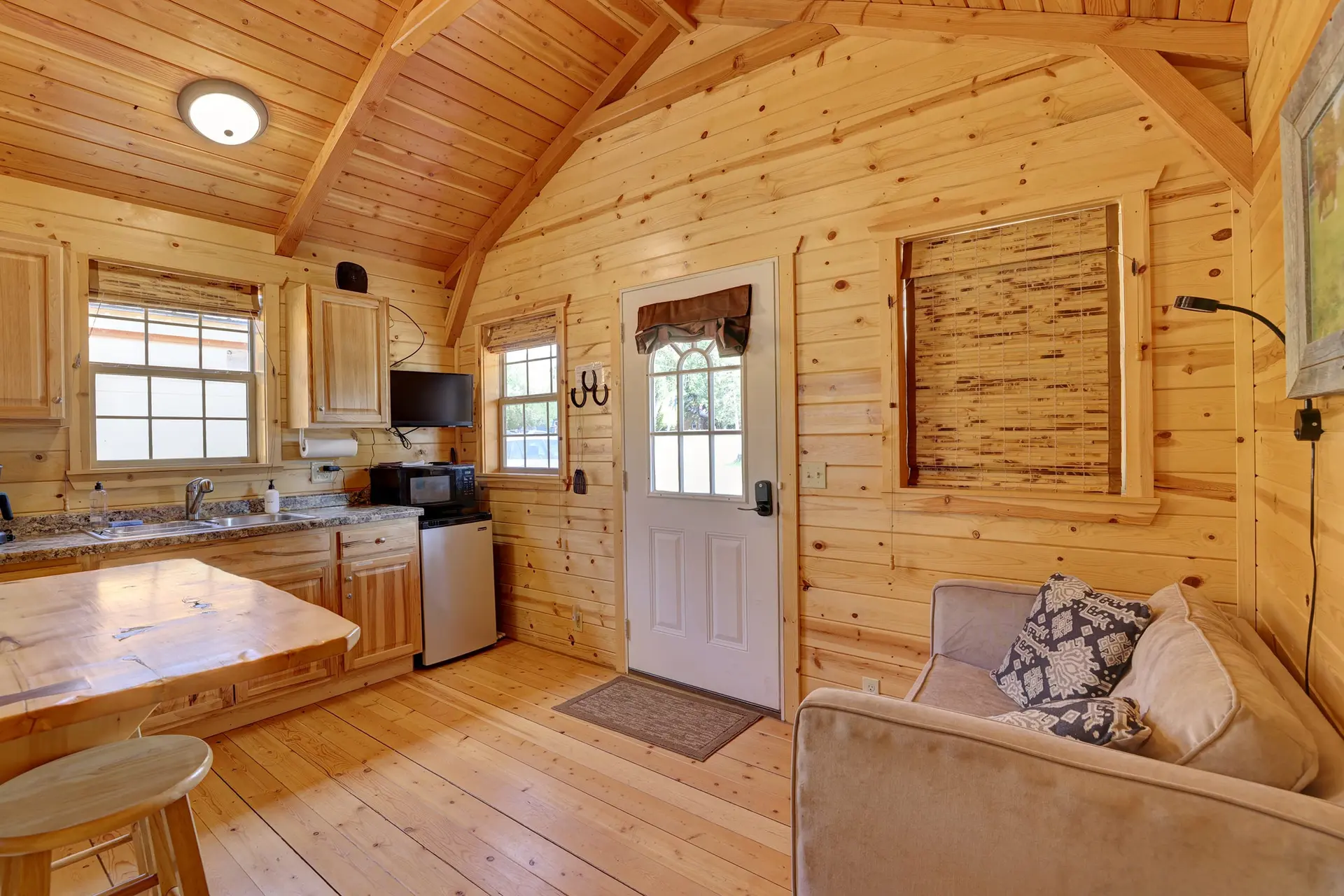 A cozy cabin interior with wood-paneled walls and ceiling, a kitchen area, a couch, and a door.