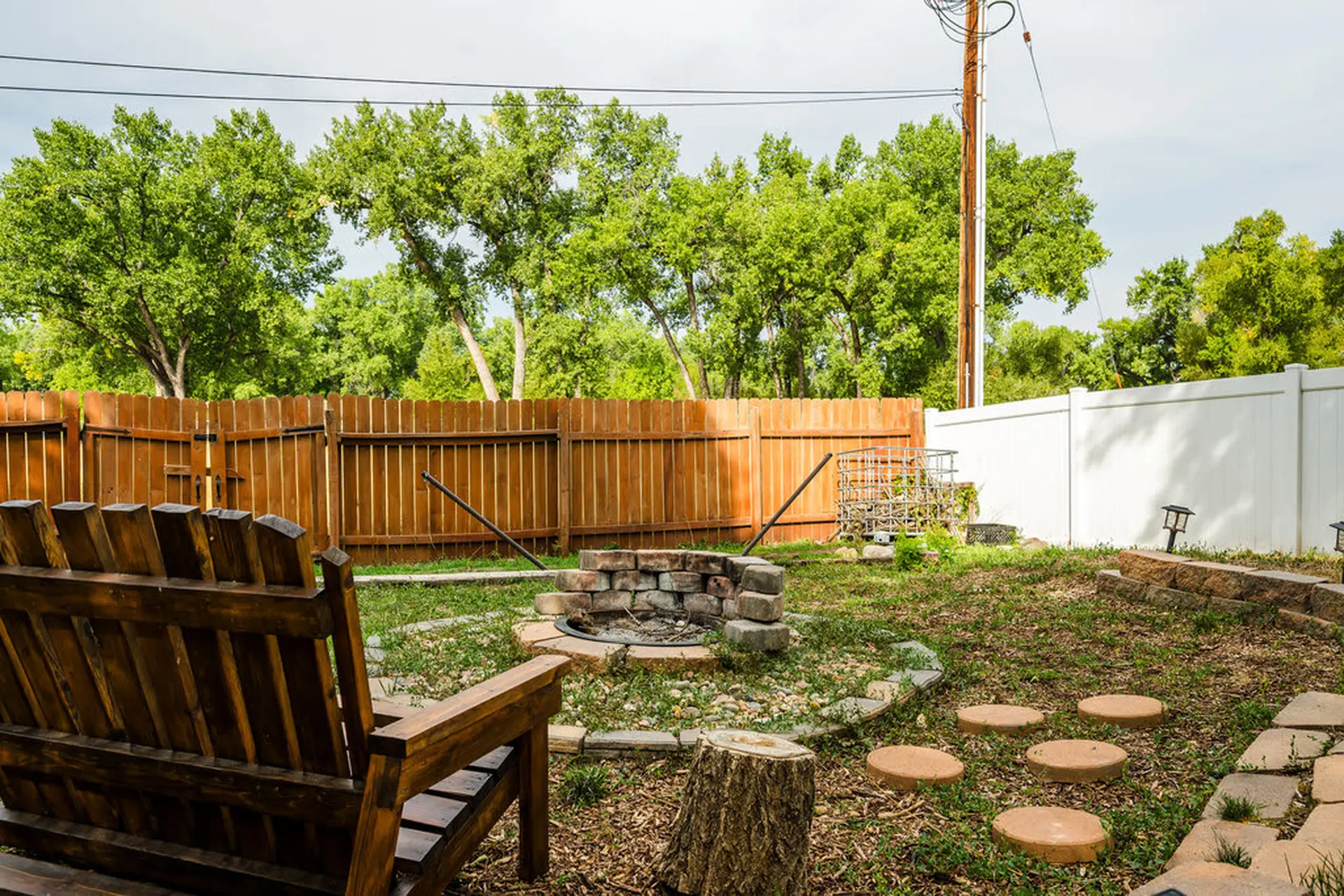 A wooden bench sits in a grassy backyard with a stone fire pit and stepping stones.