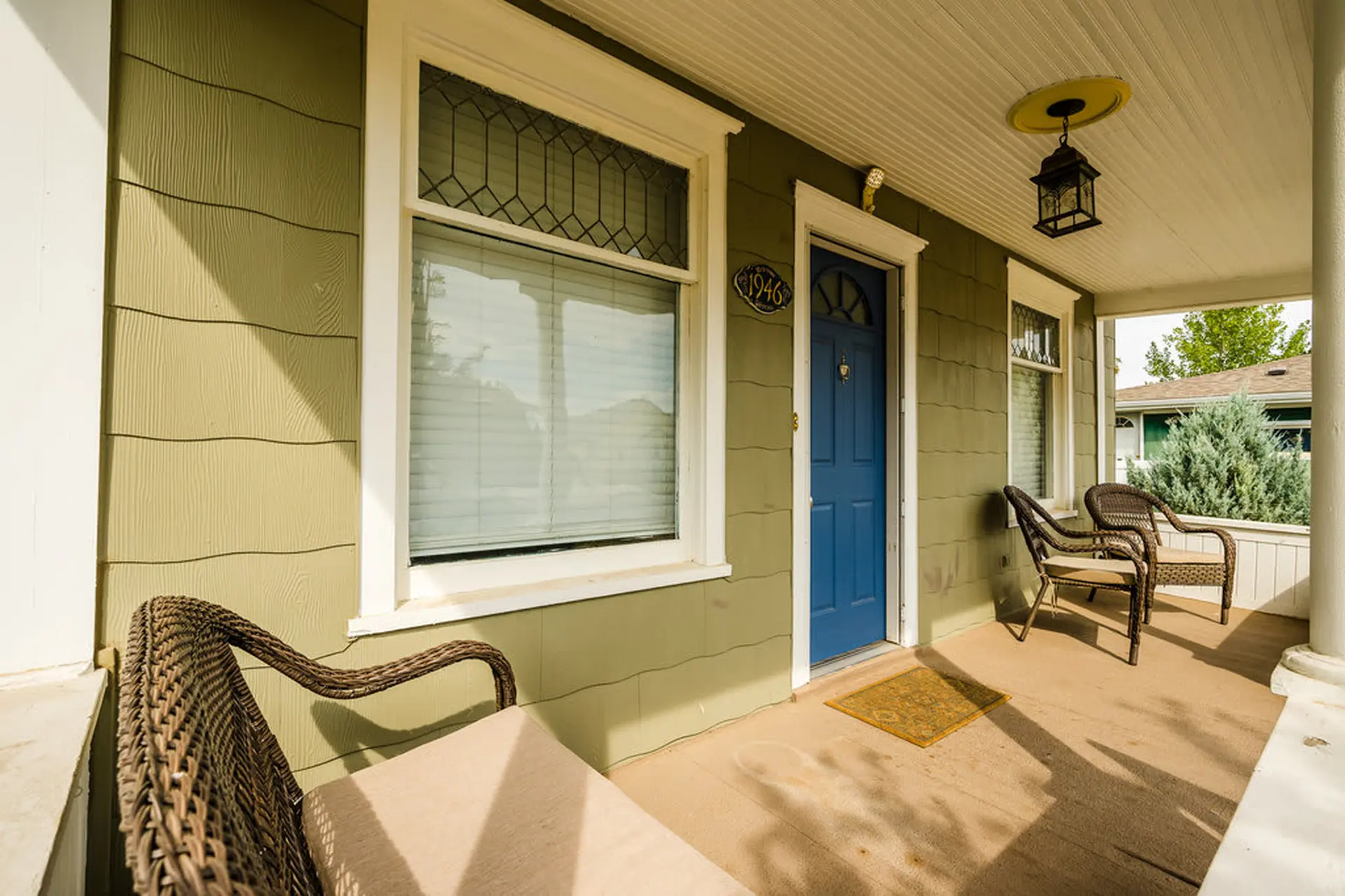 The front porch of a house has green siding, a blue door, and wicker chairs.