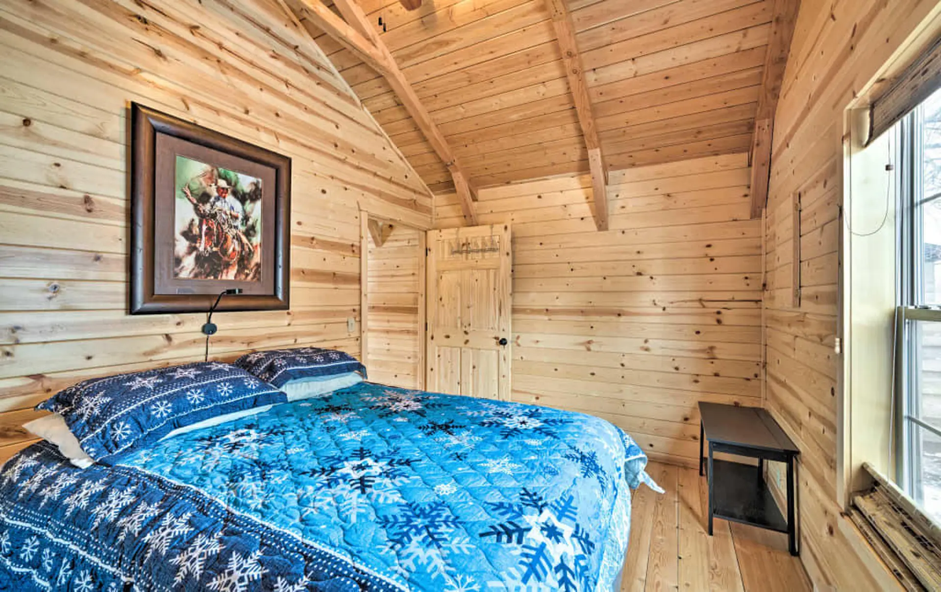 A cozy bedroom with a wooden interior, a queen bed, and a painting of a cowboy.