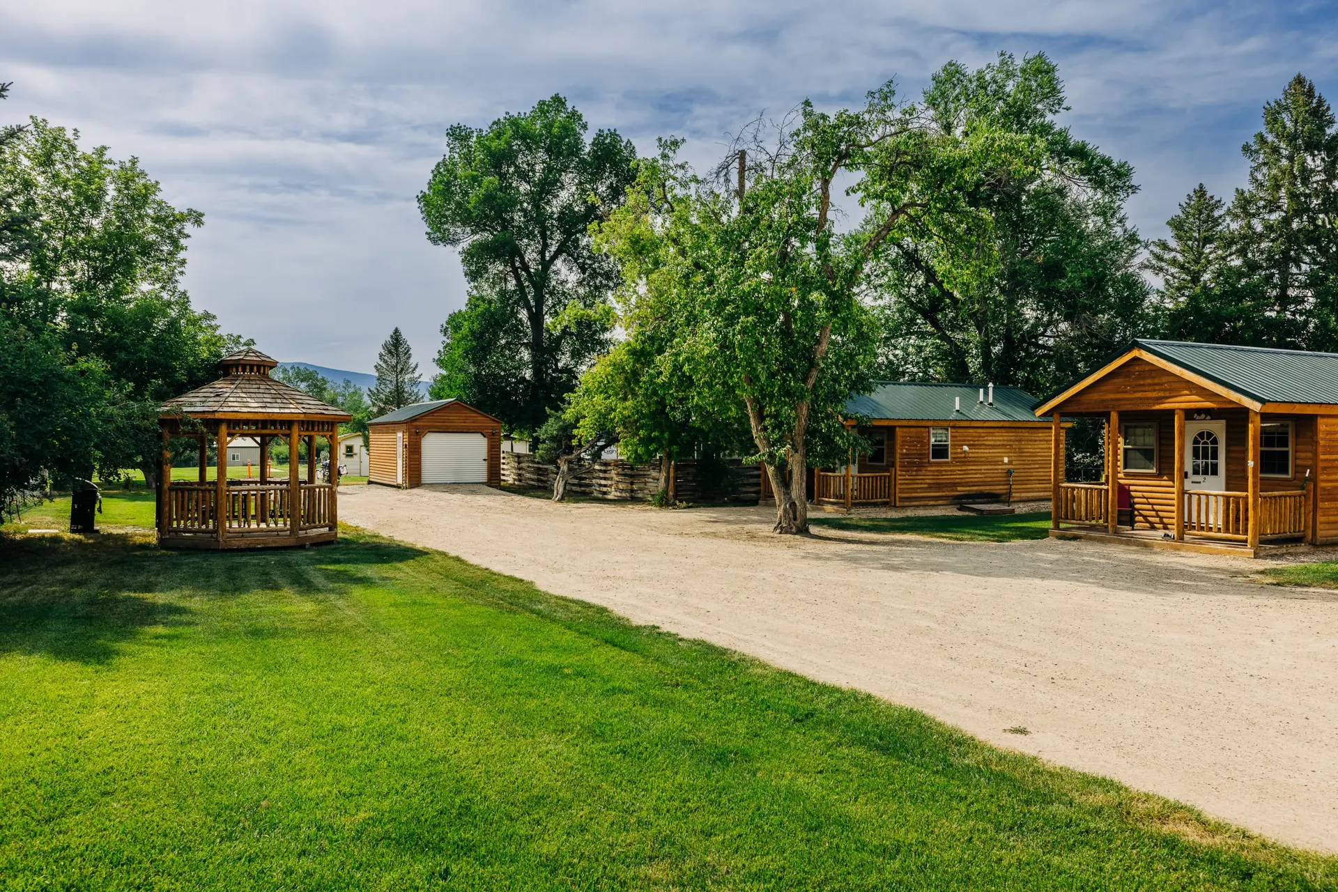 A wooden gazebo and cabins sit on a dirt road bordered by a lush green lawn and tall trees.