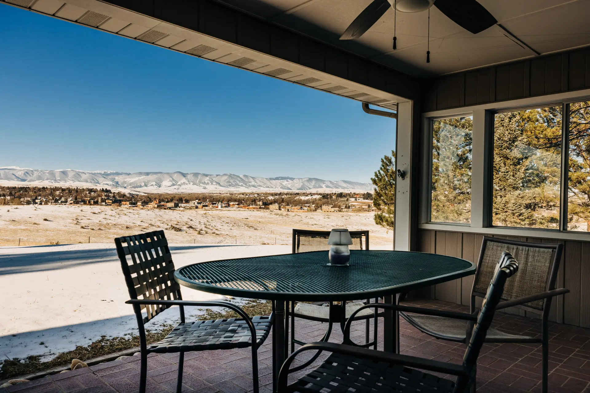 A covered patio with a table and chairs overlooks a snowy landscape with mountains.