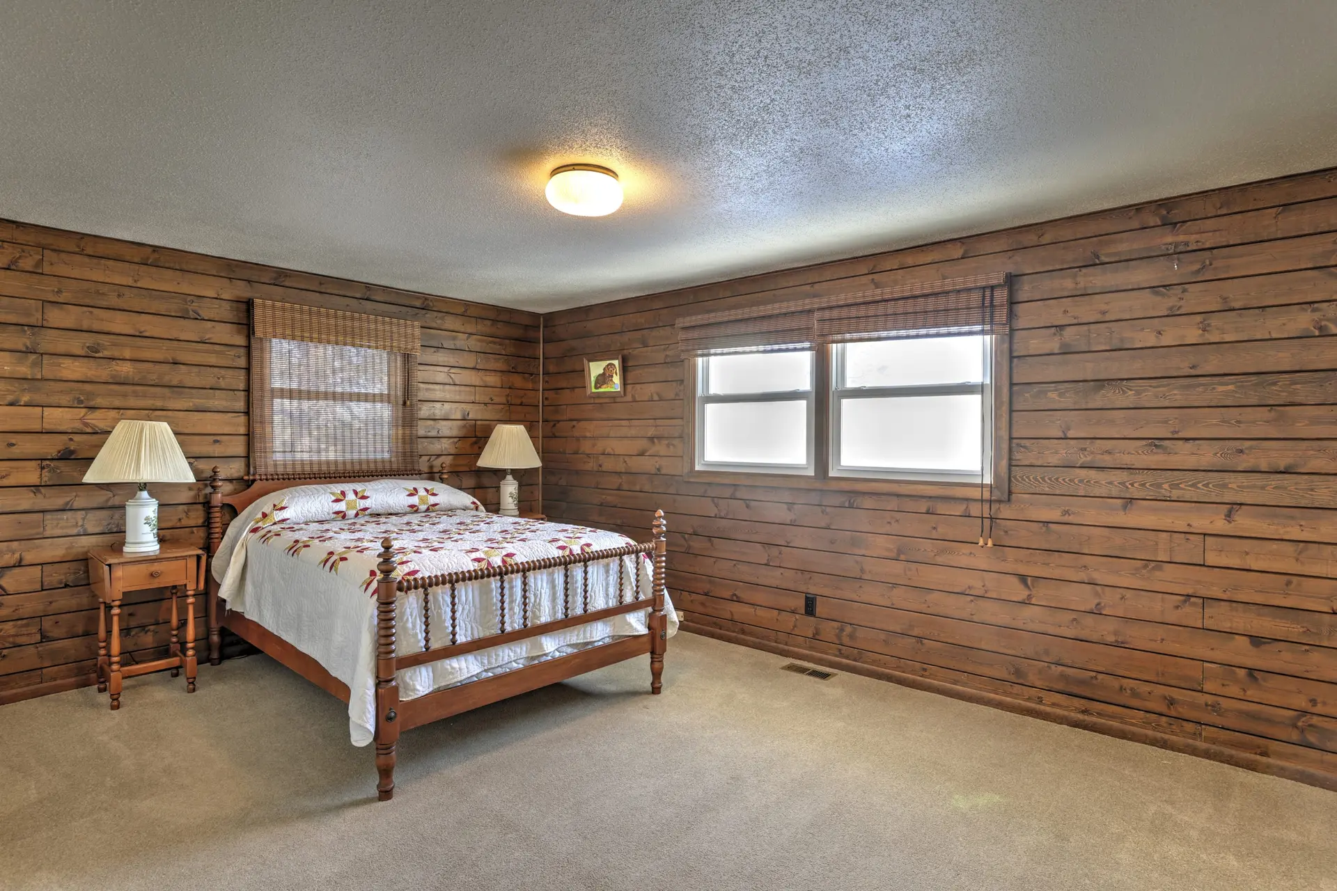 A rustic bedroom features wood-paneled walls, a wooden bed with a patterned quilt, and two windows w