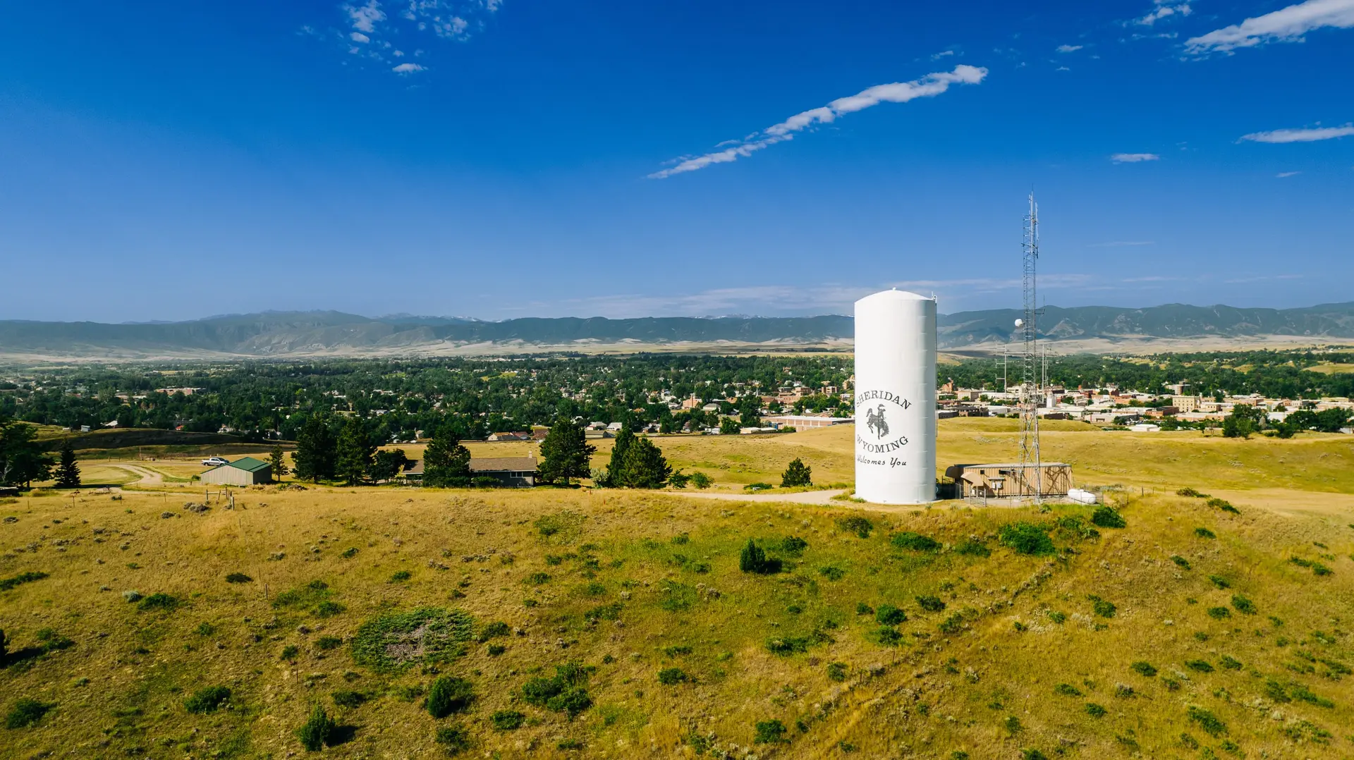 A white water tower with "SHERIDAN WYOMING Welcomes You" written on it stands on a hill overlooking 