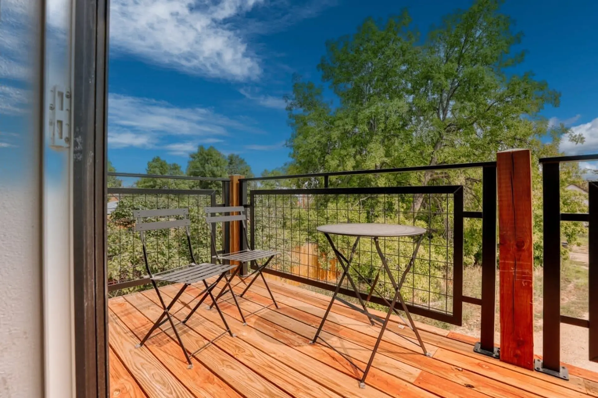 A balcony with a wooden floor, a table and chairs, and a view of trees and a blue sky.