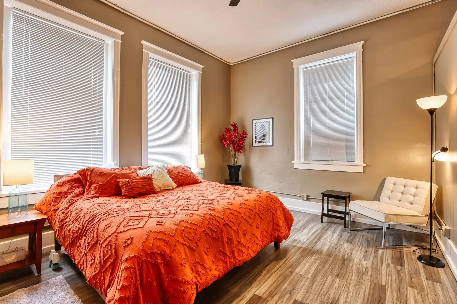 Bedroom with an orange comforter, two windows with blinds, and a chair.