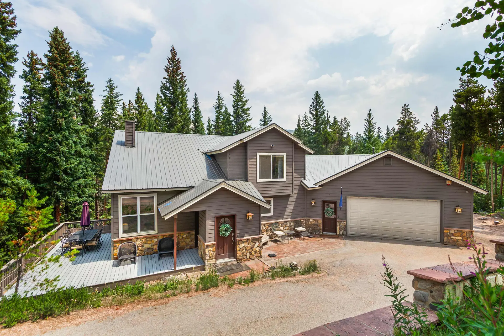 A brown house with a metal roof, a deck, and a two-car garage in a wooded area.